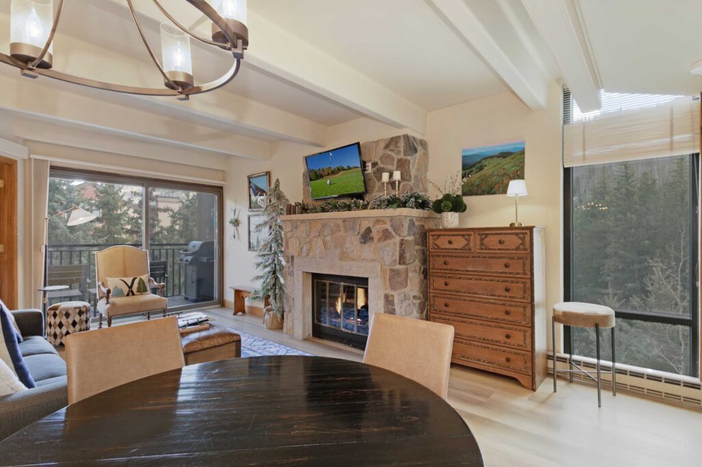 Living room of Antlers at Vail condo 502 showcasing a lit stone fireplace, wall-mounted TV, exposed beam ceiling, and sliding glass doors to the balcony