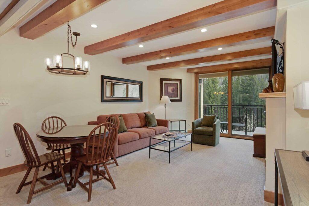 Antlers at Vail condo 504 open-concept living and dining area featuring a gas fireplace with gold trim and flat-screen TV above, exposed wood ceiling beams, glass coffee table, round dining table with Windsor chairs, kitchen bar seating, and a chandelier