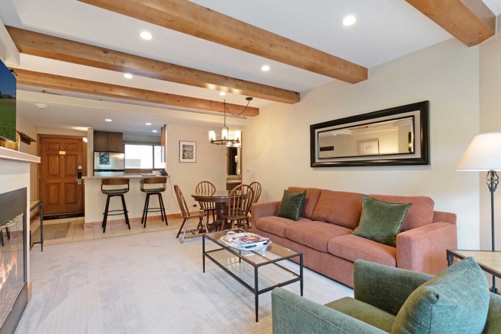 Antlers at Vail condo 504 open-concept living and dining area featuring a gas fireplace with gold trim and flat-screen TV above, exposed wood ceiling beams, glass coffee table, round dining table with Windsor chairs, kitchen bar seating, and a chandelier