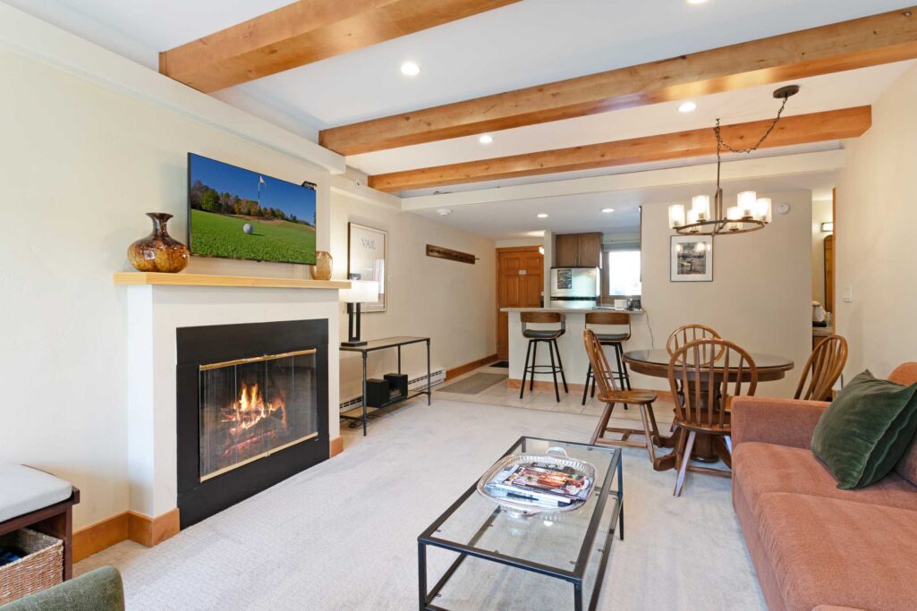 Antlers at Vail condo 504 open-concept living and dining area featuring a gas fireplace with gold trim and flat-screen TV above, exposed wood ceiling beams, glass coffee table, round dining table with Windsor chairs, kitchen bar seating, and a chandelier