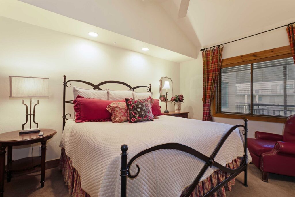 Antlers at Vail condo 515 primary bedroom with ornate iron bed frame, red and floral decorative pillows, plaid curtains, vaulted ceiling with recessed lighting, and red leather accent chair