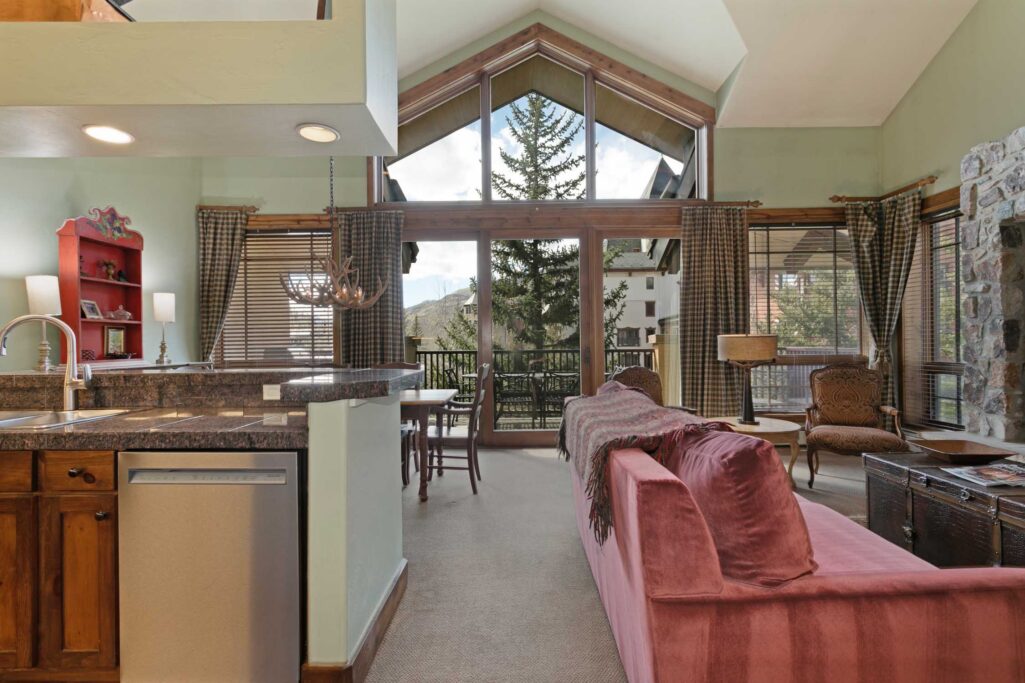 Antlers at Vail condo 515 open-concept living area viewed from kitchen showing granite island, pink sofa with throw blanket, antler chandelier, dining table, and floor-to-ceiling triangular windows with mountain and pine tree views