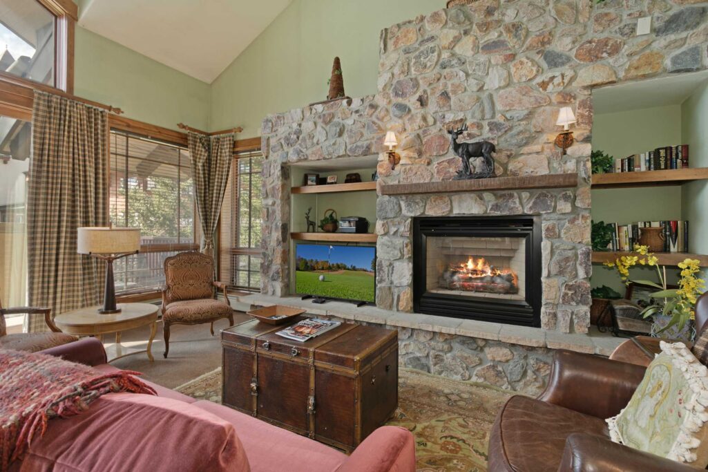 Antlers at Vail condo 515 living room with dramatic floor-to-ceiling stone fireplace, built-in shelving, flat-screen TV, bronze deer sculpture on mantel, vintage trunk coffee table, pink sofa, plaid curtains, and vaulted ceiling