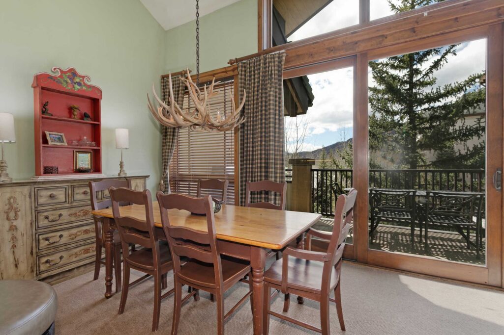 Antlers at Vail condo 515 dining area with pine farm table, six wood chairs, antler chandelier, painted red hutch, distressed sideboard, and sliding glass door opening to balcony with mountain and pine tree views