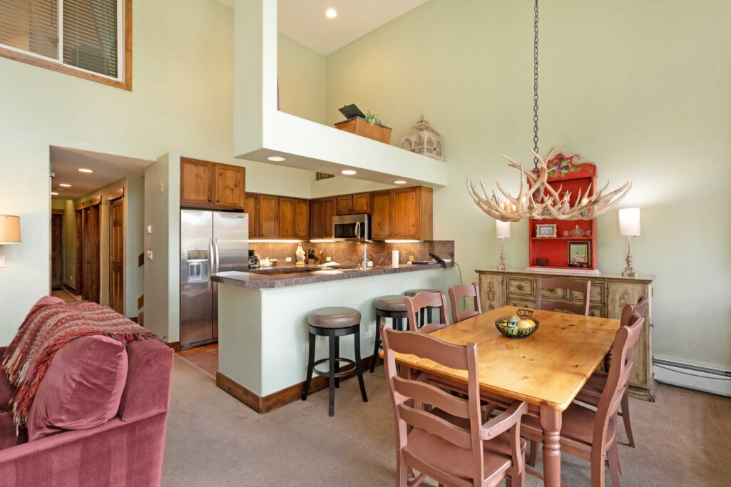 Antlers at Vail condo 515 open-concept dining and kitchen area with antler chandelier, pine dining table, bar seating at granite island, stainless steel appliances, warm wood cabinetry, and two-story vaulted ceiling