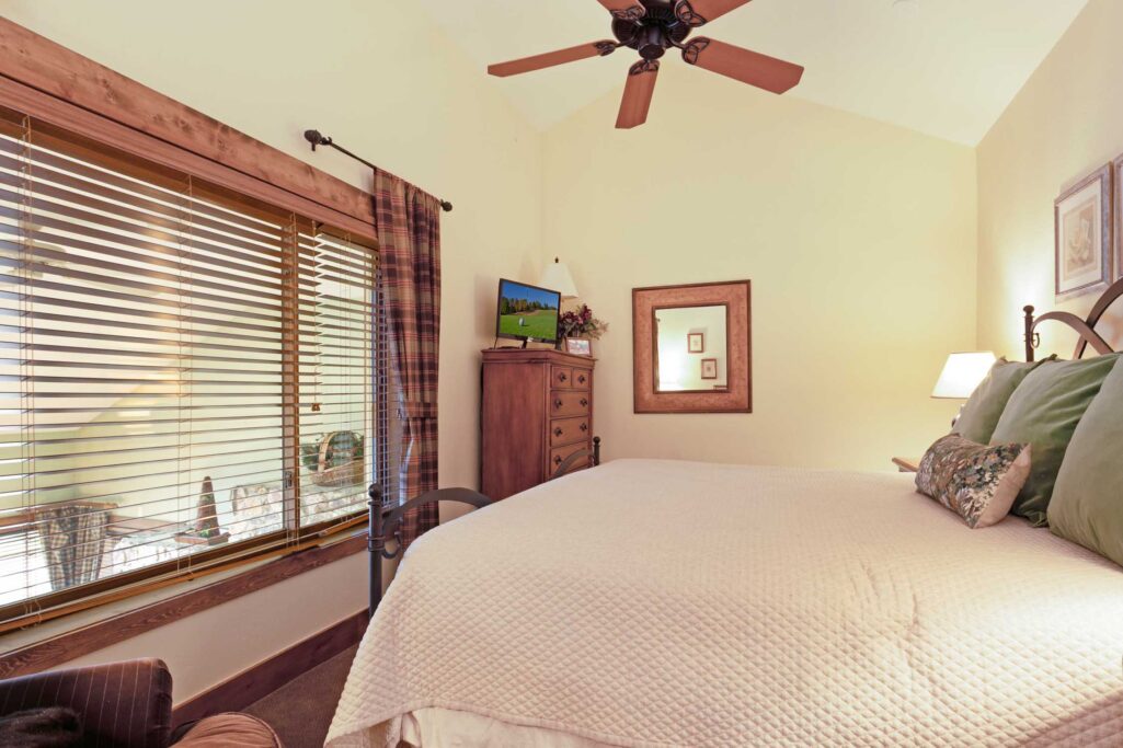 Antlers at Vail condo 515 bedroom with iron bed frame, green accent pillows, wood dresser with flat-screen TV, framed mirror, ceiling fan, and large wood-framed window with plaid curtains