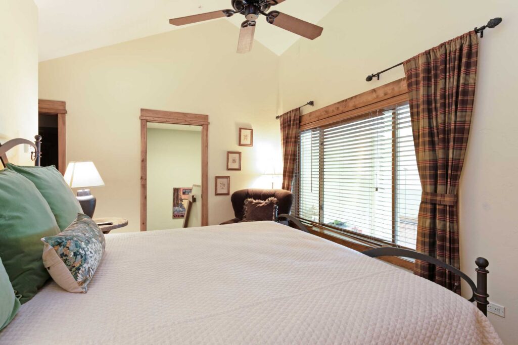 Antlers at Vail condo 515 bedroom alternate view with iron bed frame, green and floral pillows, ceiling fan, plaid curtains, large window, dark accent chair, and doorway to hallway with small framed artwork gallery wall