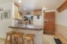 Antlers at Vail condo 607 kitchen bar seating area with three rustic log stools at granite breakfast bar, wood cabinetry, white refrigerator, and wood entry door with keypad lock visible alongside a log bench