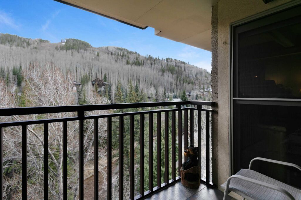 Antlers at Vail condo 607 private balcony with black metal railing, outdoor seating, decorative bear sculpture, and panoramic views of Vail mountain with aspen and pine trees under blue sky
