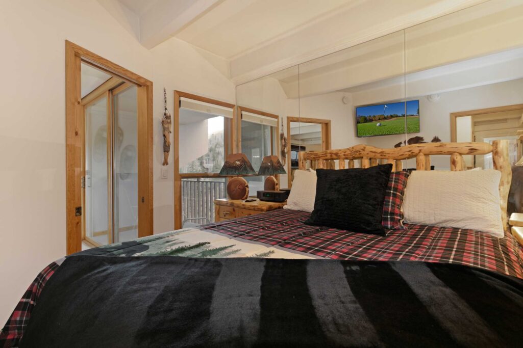 Antlers at Vail condo 607 primary bedroom alternate view showing log bed frame, black and plaid bedding, mountain-themed lamp, wood nightstand, mirrored wall, flat-screen TV, and sliding glass door to private balcony with tree views