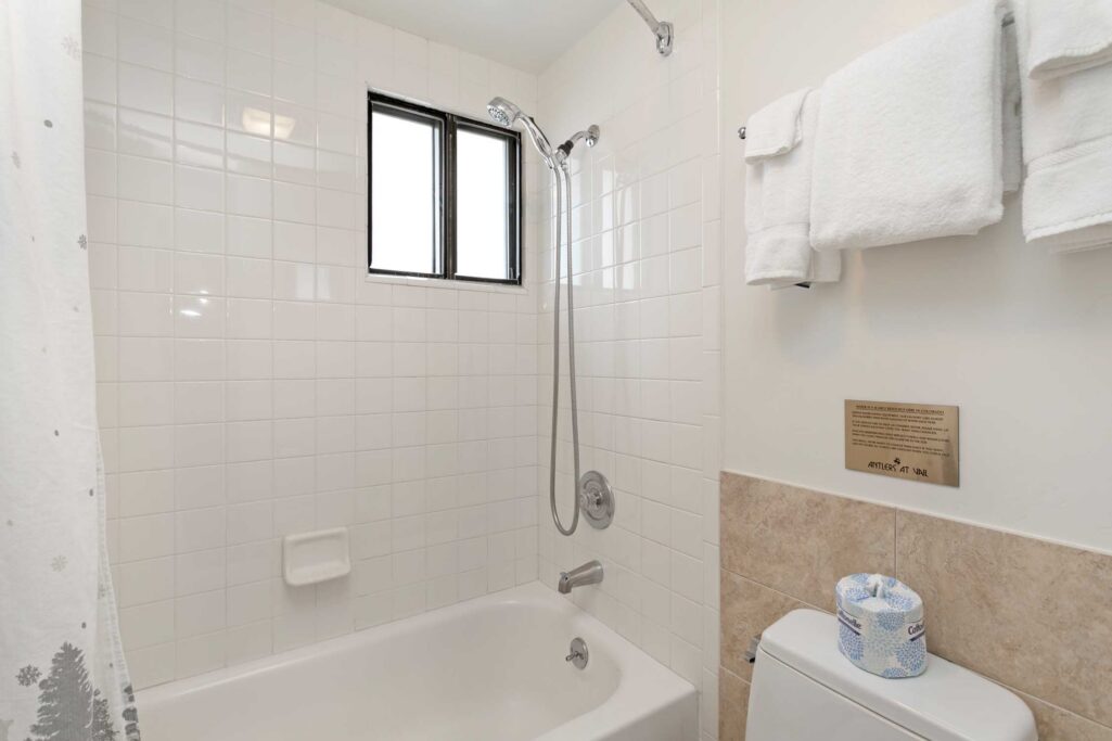 Antlers at Vail condo 607 tub-shower bathroom with white subway tile surround, handheld showerhead, small window, white towels on rack, beige tile wainscoting, and Antlers at Vail water conservation placard